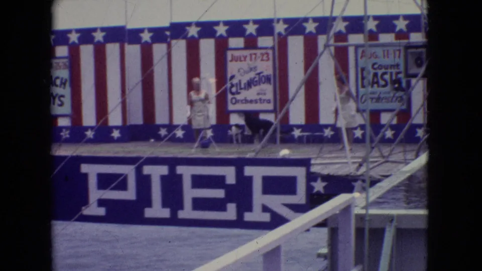 1965 home-movie frame of the Steel Pier stage showing performers in light-colored clothing, a patriotic backdrop, partial orchestra posters, and pier structure below.