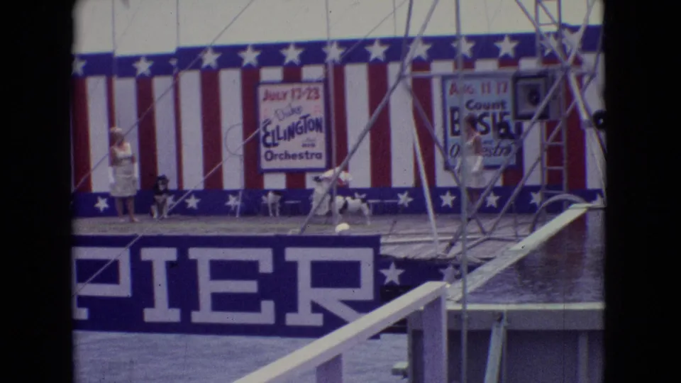 1965 Atlantic City Steel Pier stage view with a woman in a light dress, a dog near the backdrop, visible Duke Ellington and Count Basie posters, and large pier signage below.