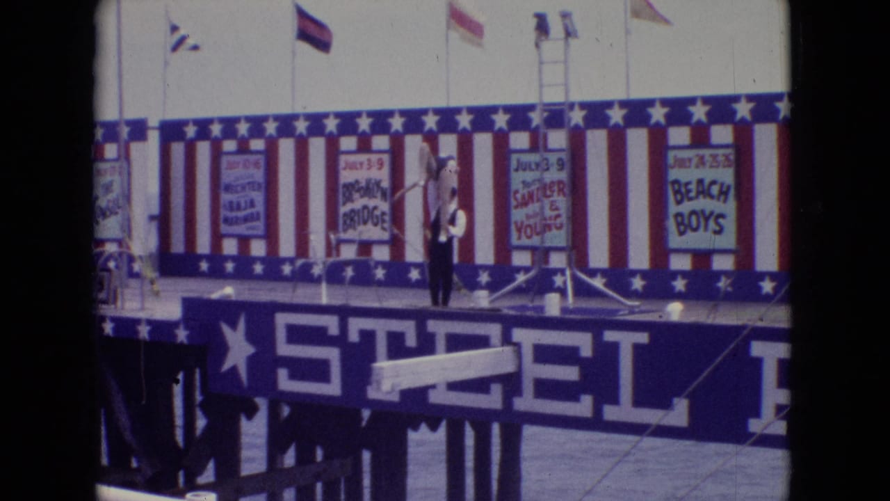 1965 New Jersey 8mm frame showing a male acrobat in dark clothing lifting a female performer in white on a Steel Pier stage with striped backdrop and event posters.