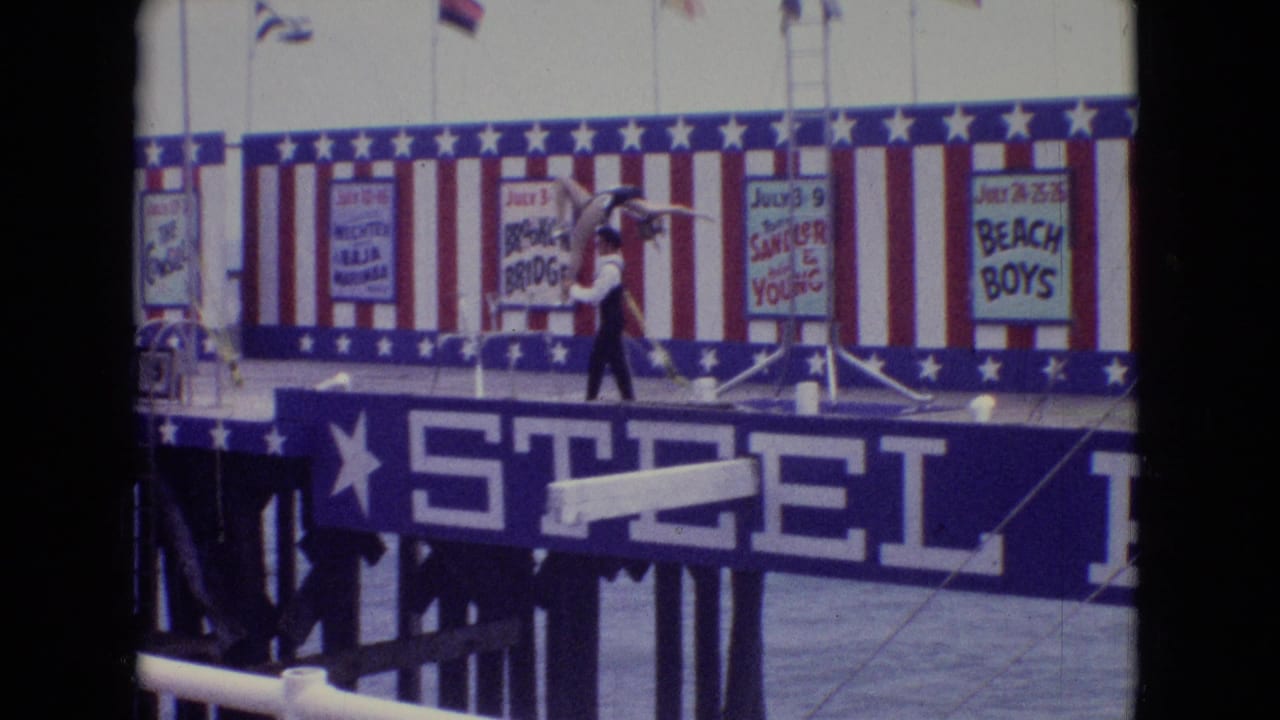 1965 New Jersey frame of the Steel Pier act with the male performer moving across the stage while supporting the female performer near posters and patriotic bunting.