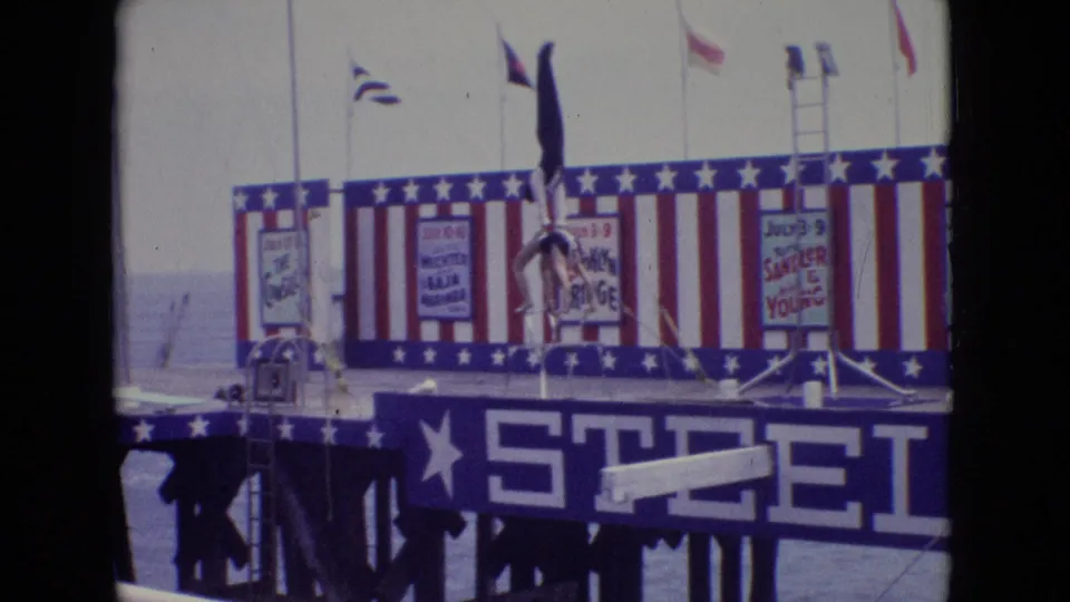 1965 New Jersey frame of the acrobatic handstand held in front of the patriotic Steel Pier stage backdrop, with event posters and flags visible behind the performers.