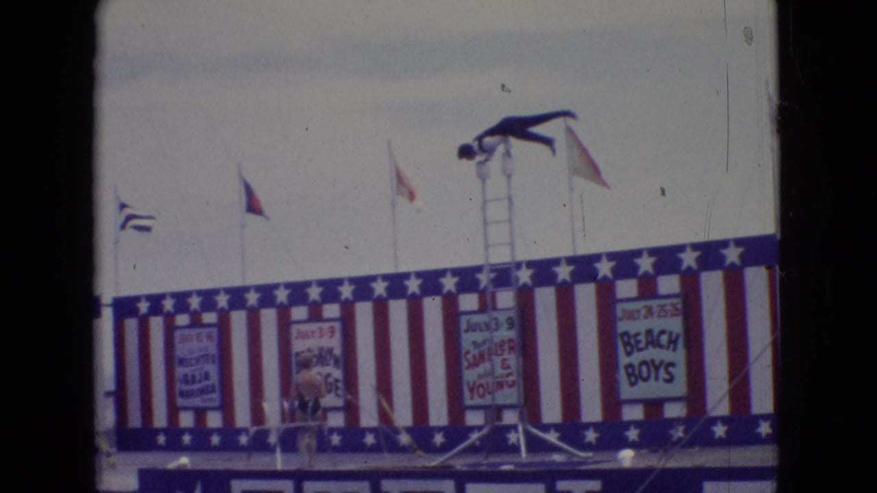 In 1965 New Jersey home-movie footage, a diver in dark trunks balances atop a tall ladder over a red, white, and blue pier stage with flags and painted show signs.