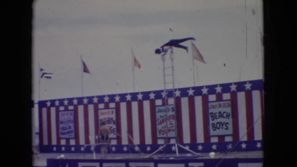 In 1965 New Jersey home-movie footage, a diver in dark trunks balances atop a tall ladder over a red, white, and blue pier stage with flags and painted show signs.