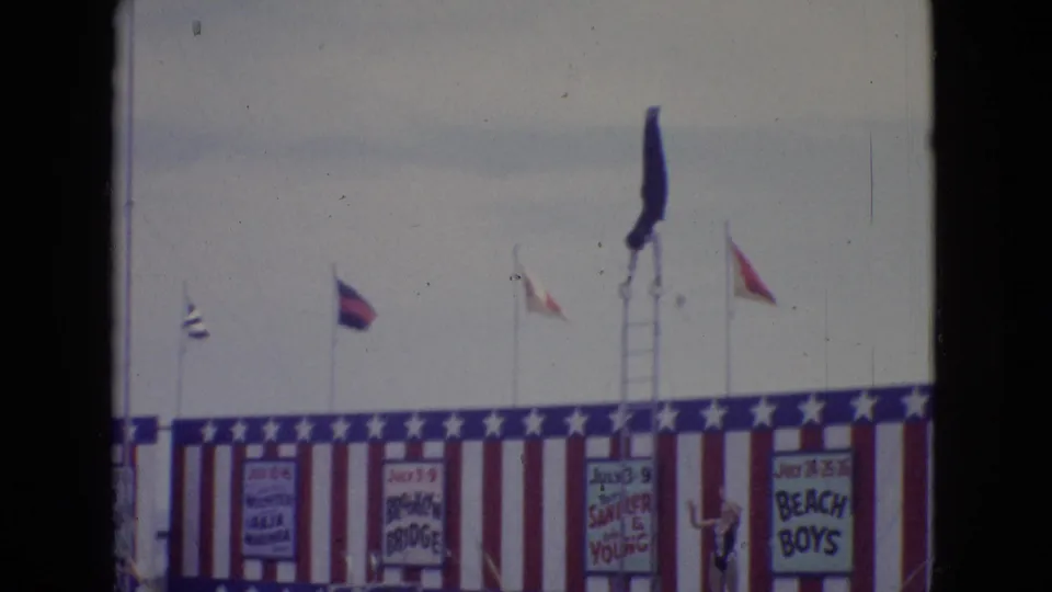 A patriotic amusement-pier stage in New Jersey shows flags, hand-painted signs, and a tall ladder with a performer high above the platform.