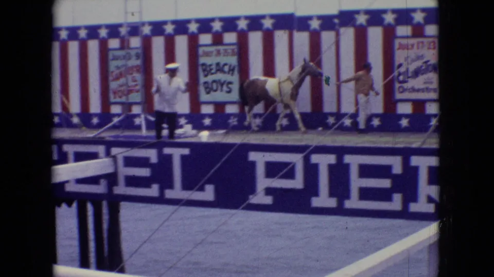 A small horse is led across the red, white, and blue stage as performers stand nearby, with pier lettering and painted act signs visible in the 1965 New Jersey footage.