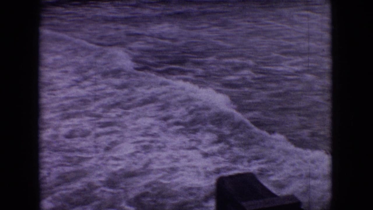 1965 New Jersey 8mm frame showing a foamy wave rolling across dark rippled shoreline water, with a dark structure partly visible near the lower edge.