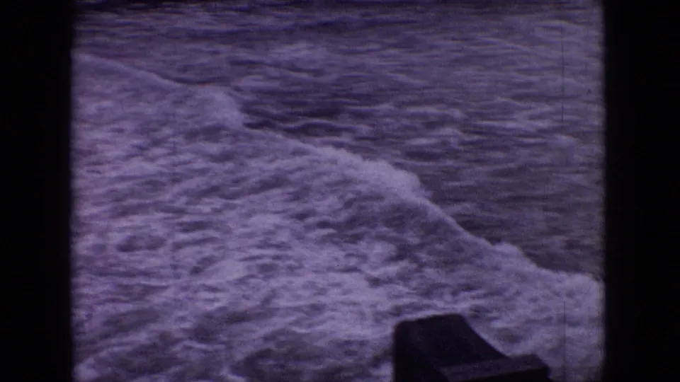 1965 New Jersey 8mm frame showing a foamy wave rolling across dark rippled shoreline water, with a dark structure partly visible near the lower edge.