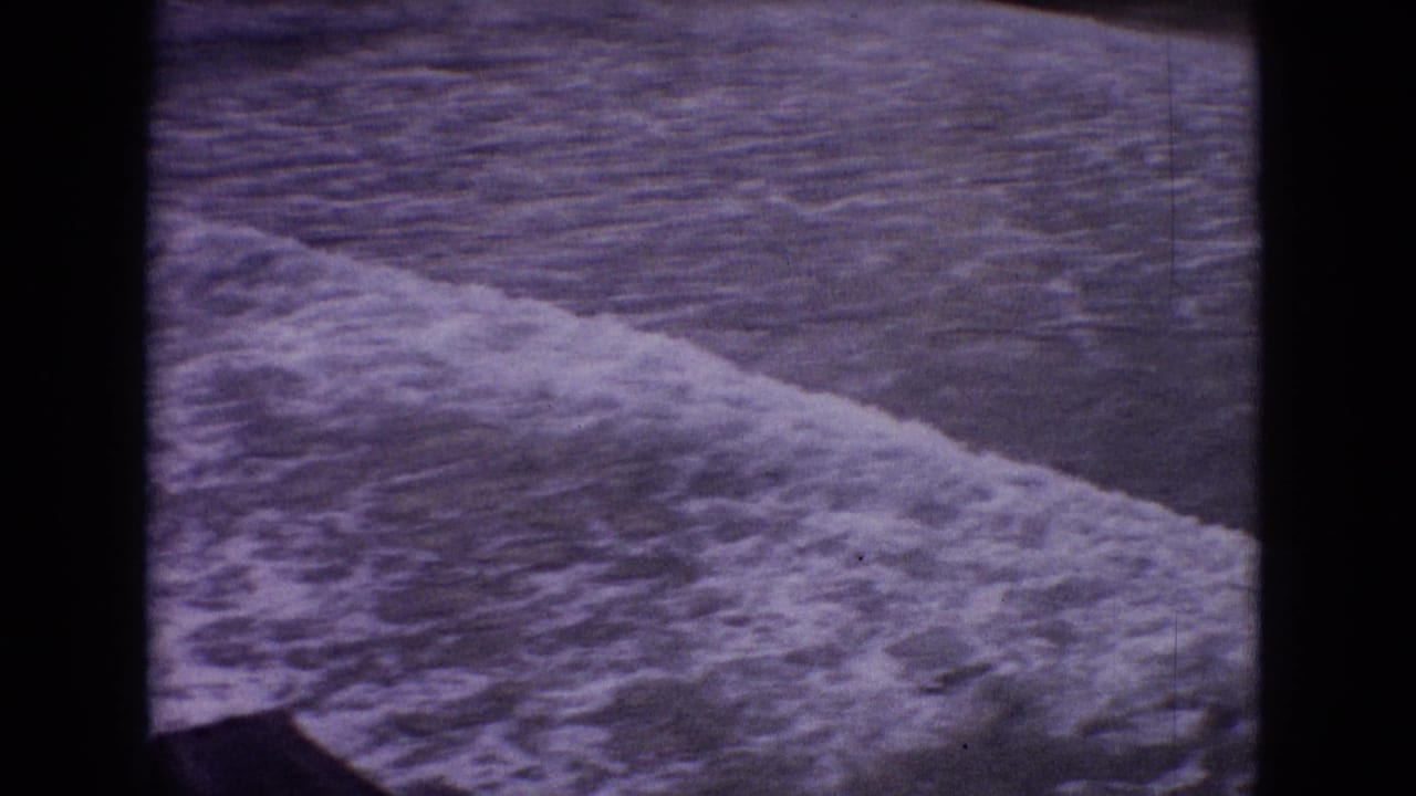1965 New Jersey 8mm frame with white surf moving diagonally across rippled water, bordered by dark film-edge shadows.