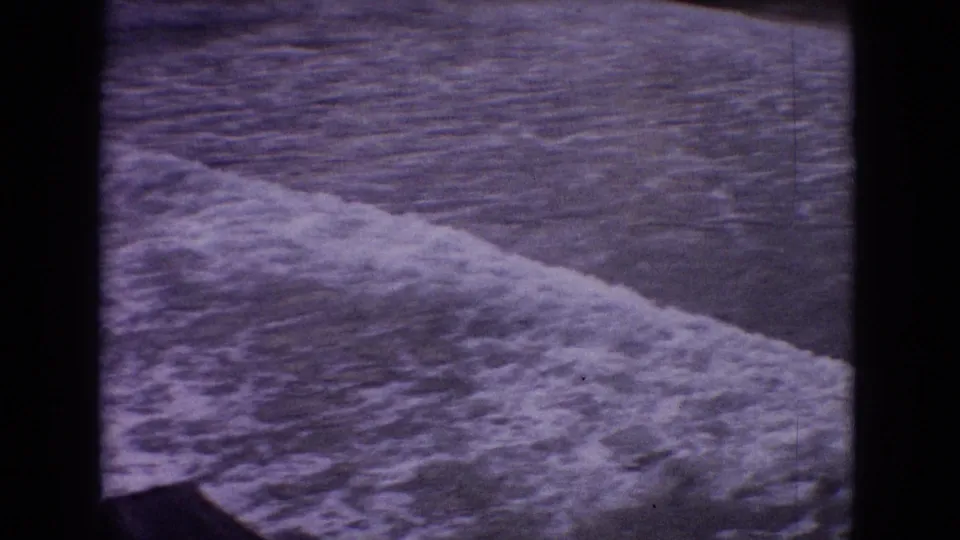 1965 New Jersey 8mm frame with white surf moving diagonally across rippled water, bordered by dark film-edge shadows.
