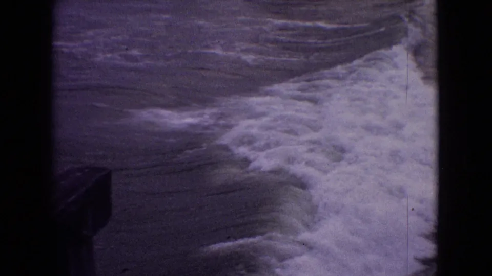 1965 New Jersey 8mm frame showing foamy shoreline water spreading through the lower frame, with a dark structure clipped at the left edge.