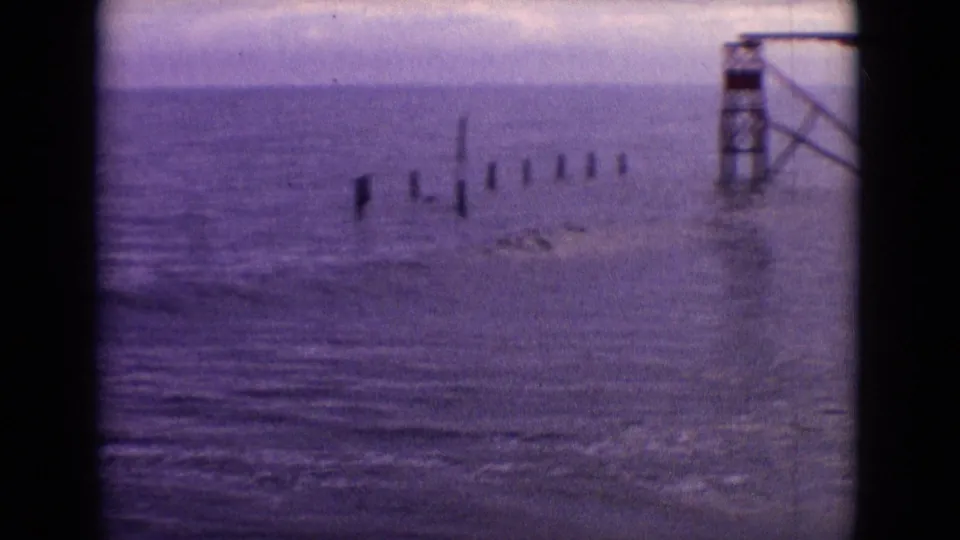 1965 New Jersey 8mm frame showing ocean water, exposed posts in the surf, and part of a weathered amusement pier structure at the right edge.