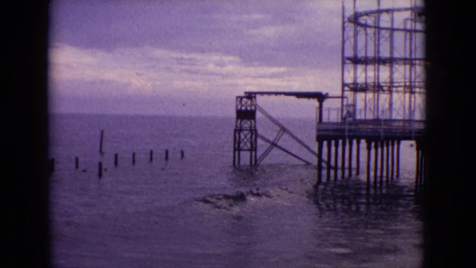1965 New Jersey 8mm frame of a weathered pier on tall pilings, with scattered posts in the ocean and waves breaking near the structure.