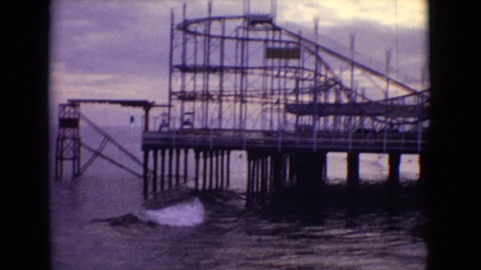 1965 New Jersey 8mm frame showing the skeletal frame of a roller coaster or amusement ride on a wooden pier, with waves breaking below the pilings.