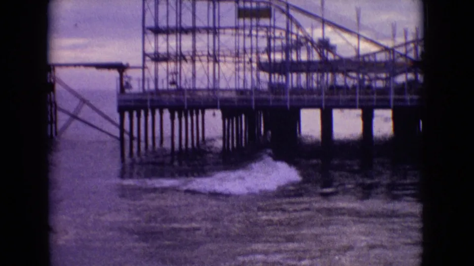 1965 New Jersey 8mm frame of an abandoned-looking amusement pier with roller coaster framework, tall pilings, and seafoam moving beneath the structure.
