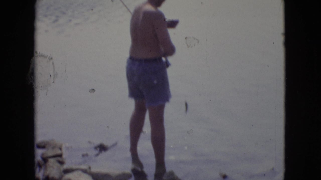 A shirtless man in blue swim trunks stands at the edge of water during a 1965 New Jersey home-movie fishing or shoreline scene.