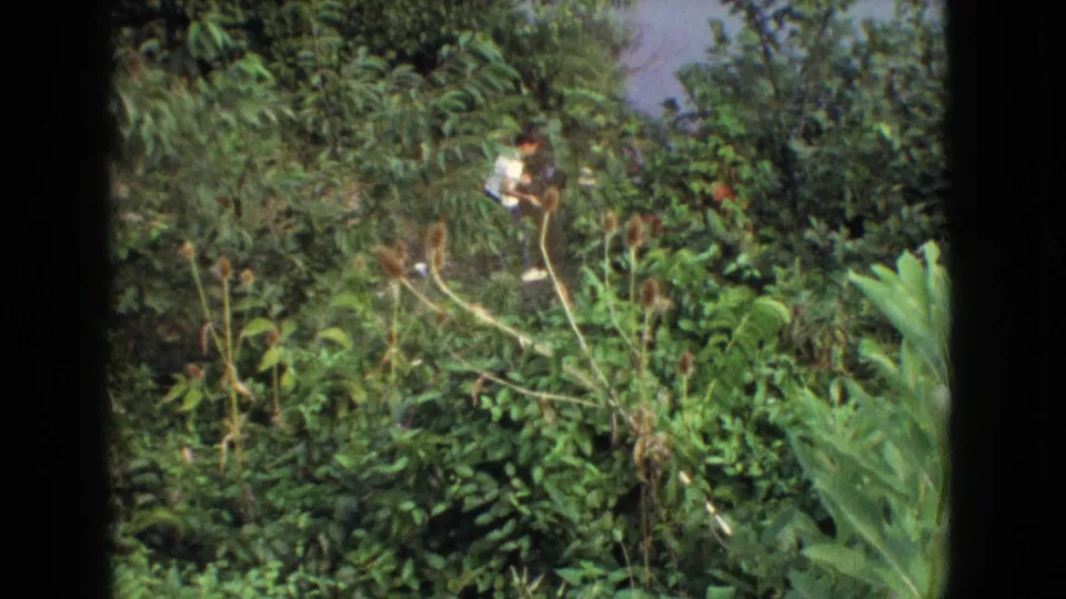 A person in a light top appears partly obscured while moving through dense green vegetation in the 1965 New Jersey home-movie scene.