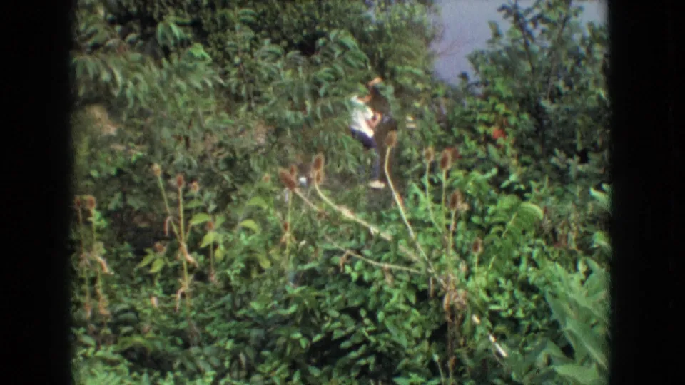 A person in a light shirt is seen among thick summer greenery, framed by the dark borders of the scanned 8mm film.