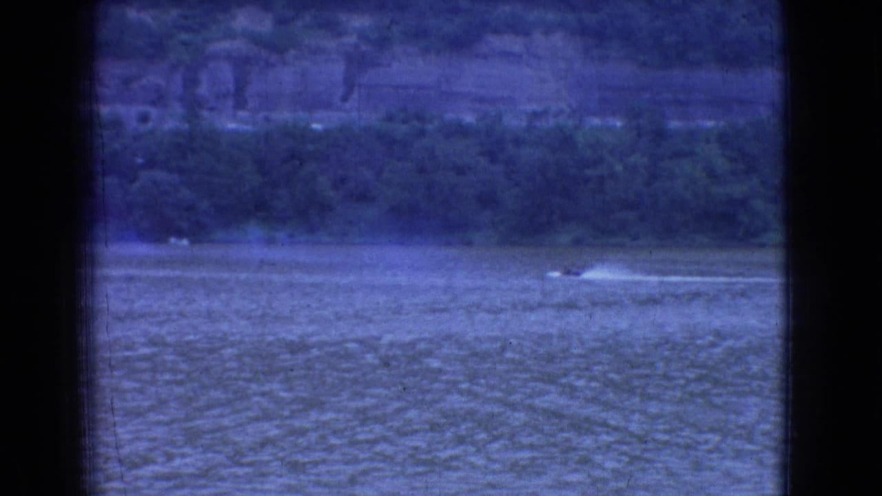 1965 New Jersey 8mm home-movie frame showing a small speedboat leaving a pale wake across a wide river below a wooded shoreline and distant cliffs.