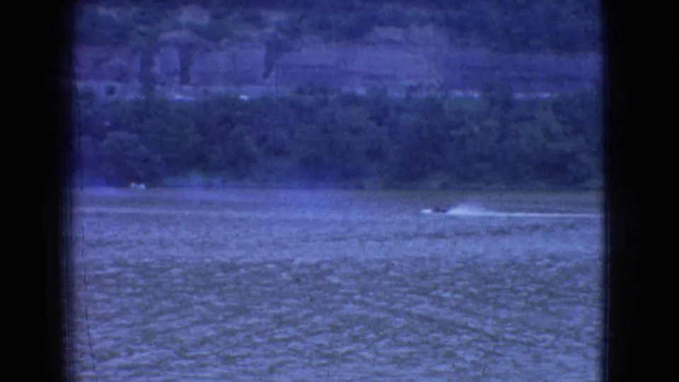 1965 New Jersey 8mm home-movie frame showing a small speedboat leaving a pale wake across a wide river below a wooded shoreline and distant cliffs.