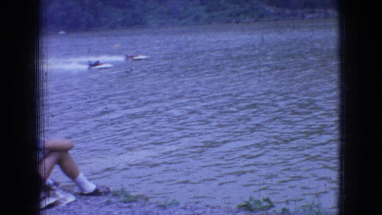 1965 New Jersey shoreline view of speedboats on rippled river water, with a seated spectator's legs partly visible in the foreground.