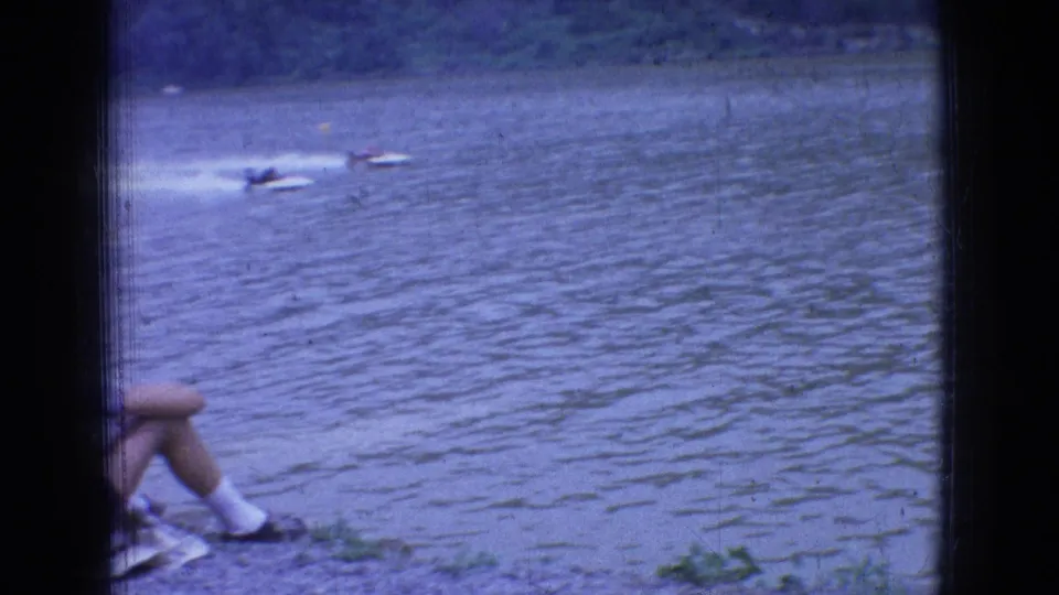 1965 New Jersey shoreline view of speedboats on rippled river water, with a seated spectator's legs partly visible in the foreground.