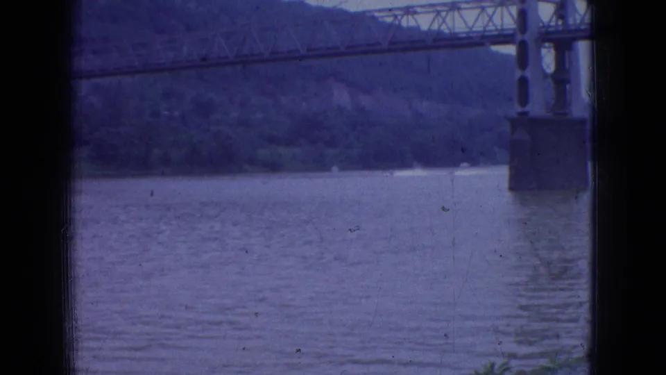 1965 New Jersey 8mm home-movie view of a wide river beneath a metal truss bridge, with wooded cliffs in the background and shoreline plants in the foreground.