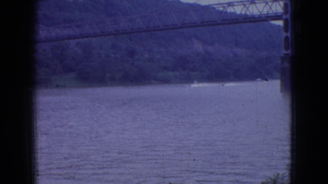 1965 New Jersey 8mm frame showing a wide river through a dark window frame, with a large suspension bridge crossing in front of wooded hills.
