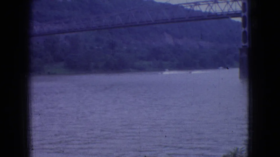 1965 New Jersey 8mm frame showing a wide river through a dark window frame, with a large suspension bridge crossing in front of wooded hills.