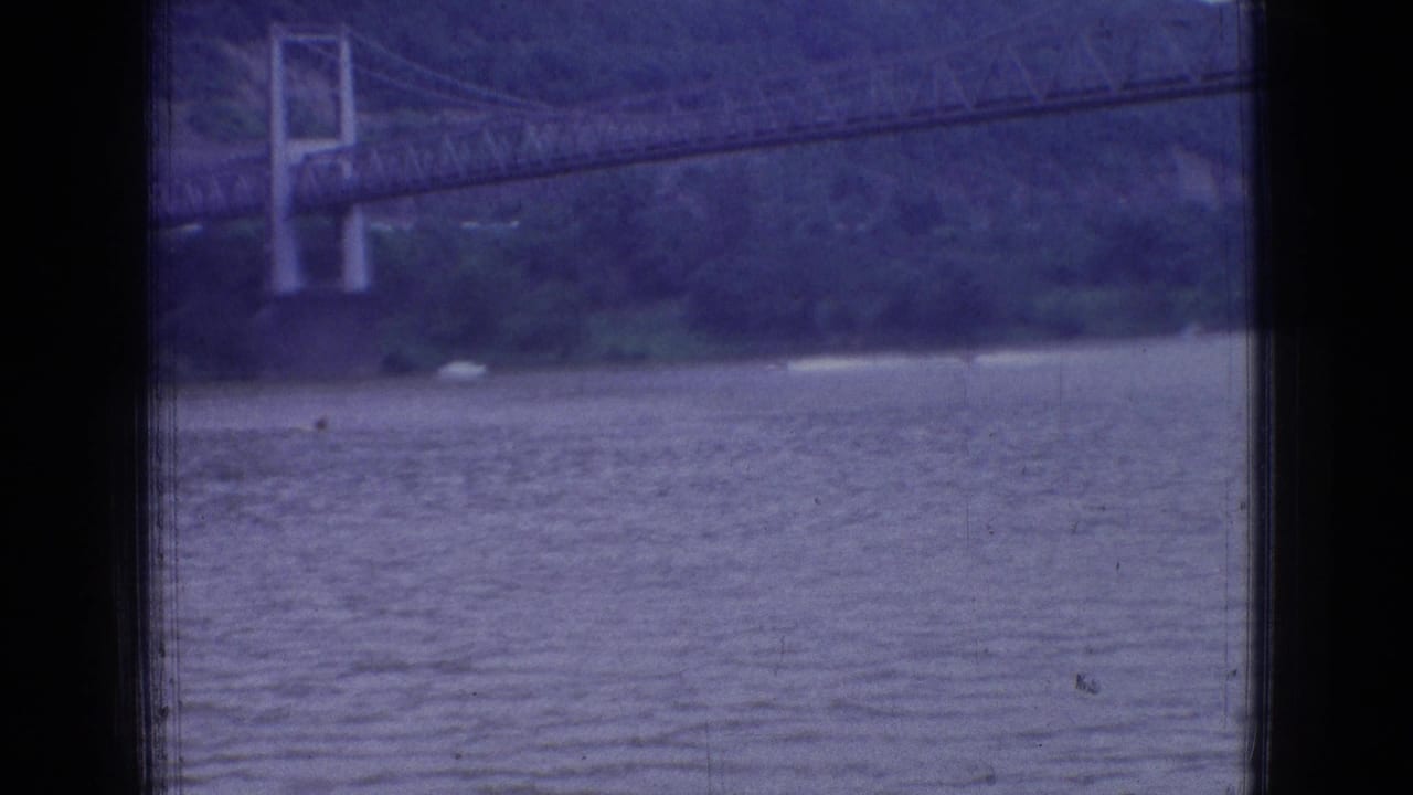 1965 New Jersey 8mm frame with the river in the foreground, a suspension bridge tower across the water, wooded shoreline, and heavy dark window framing.