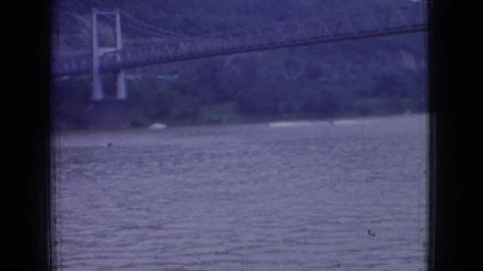 1965 New Jersey 8mm frame with the river in the foreground, a suspension bridge tower across the water, wooded shoreline, and heavy dark window framing.