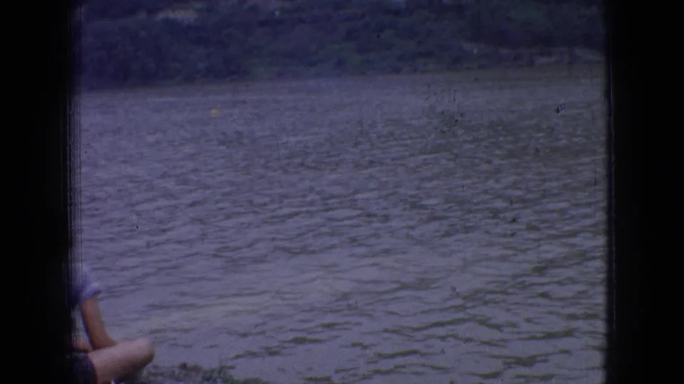 1965 New Jersey 8mm frame showing rippled river water and distant shoreline through window framing, with a partial foreground person or seat edge visible at lower left.