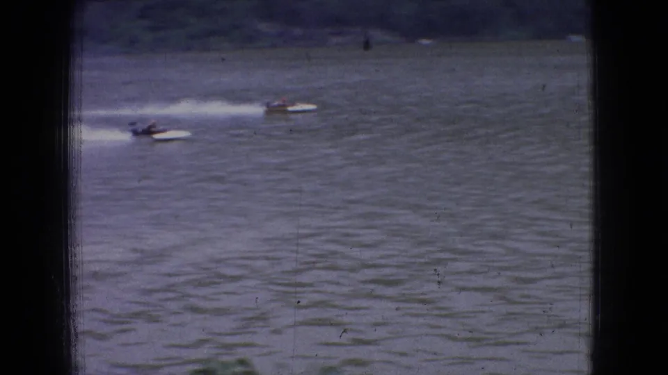 1965 New Jersey 8mm frame showing two small speedboats racing across the river below a distant suspension bridge, with white wakes trailing behind them.