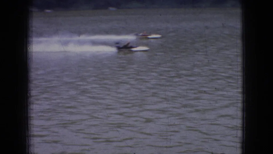 Distant powerboats speed across a wide New Jersey river in 1965 home-movie footage, leaving pale wakes on the water.