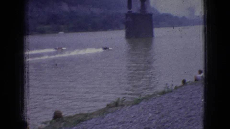Distant powerboats race along a wide river in New Jersey, with a large bridge pier or vertical structure visible near the center of the home-movie frame.