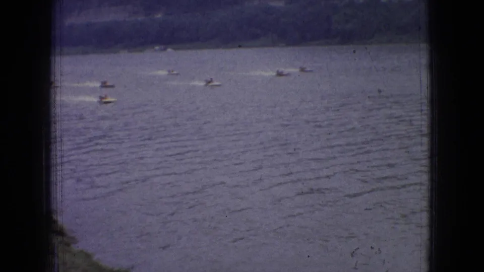 A group of small motorboats races across a wide New Jersey river in 1965 8mm footage, with distant banks and wake trails visible.