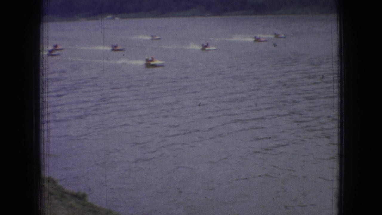 1965 New Jersey home-movie frame showing several distant small speedboats moving across rippled river water with wakes trailing behind them.