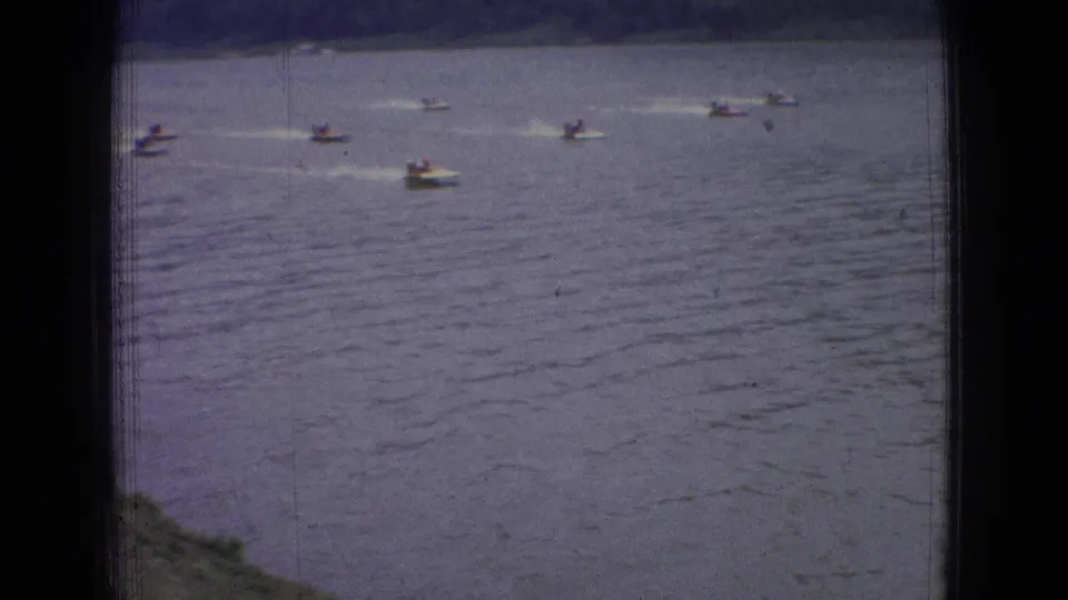 1965 New Jersey home-movie frame showing several distant small speedboats moving across rippled river water with wakes trailing behind them.