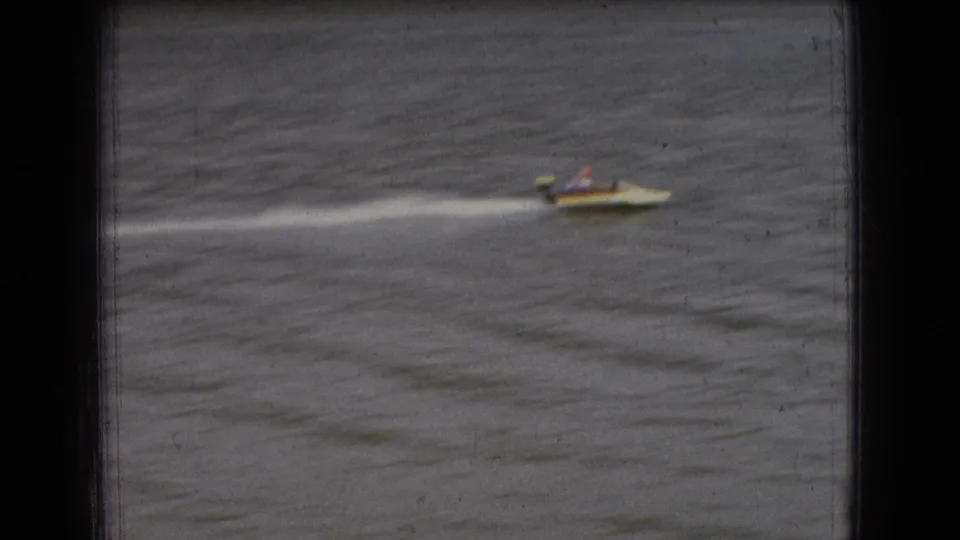 A small speedboat with tiny seated occupants races across the river, leaving a bright wake in soft color 8mm footage from the supplied 1965 New Jersey clip.