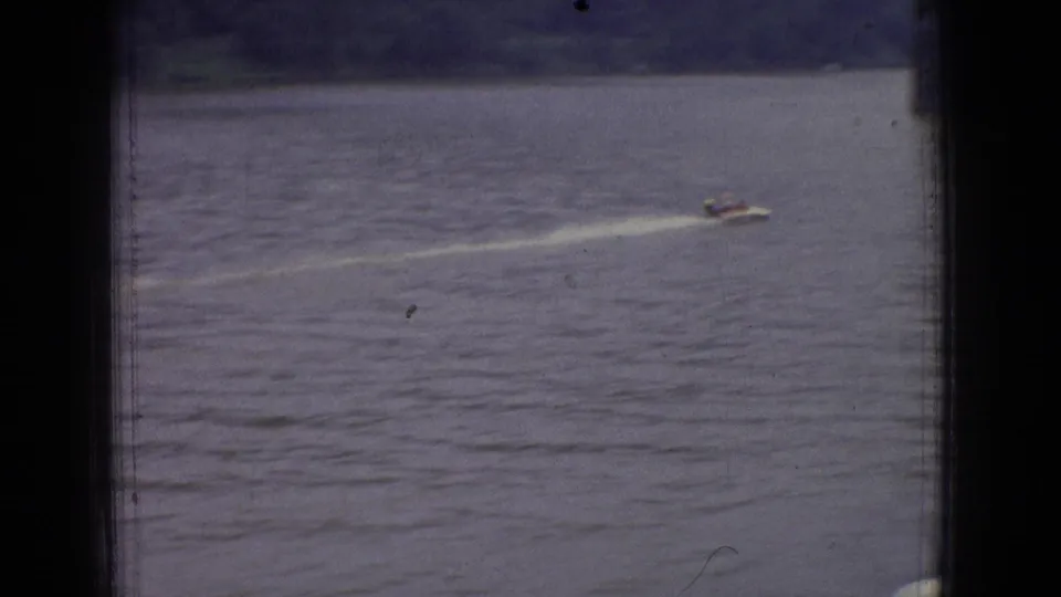 A distant small motorboat moves across open river water with a white wake, framed by dark 8mm gate edges and a faint shoreline beyond.