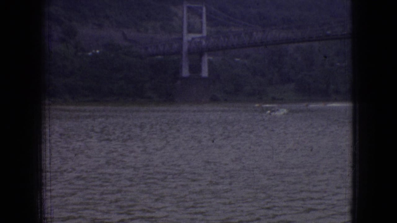 1965 New Jersey 8mm frame showing rippled river water below a distant suspension bridge tower and wooded hillside, with dark film borders visible.