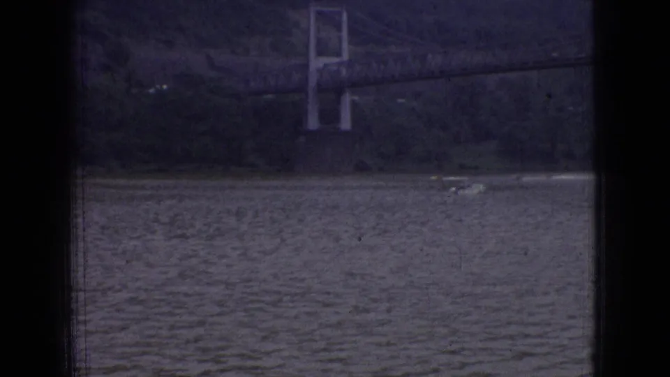 1965 New Jersey 8mm frame showing rippled river water below a distant suspension bridge tower and wooded hillside, with dark film borders visible.