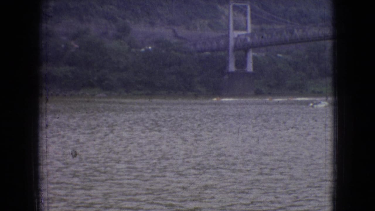 Wide 1965 New Jersey river view with a suspension bridge beyond the water and a small bright wake crossing near the distant shoreline.
