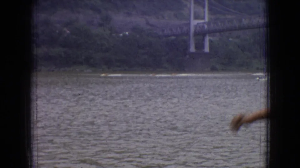 Home-movie frame of the New Jersey river and suspension bridge, partly interrupted by a blurred foreground arm or object near the lower right edge.