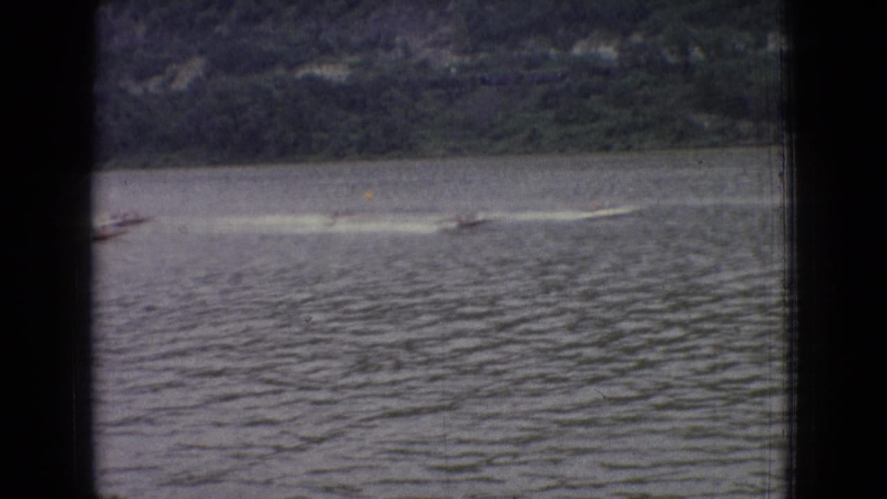 1965 New Jersey 8mm home-movie frame showing small speed boats racing across a calm lake with white wakes and a wooded far shore.