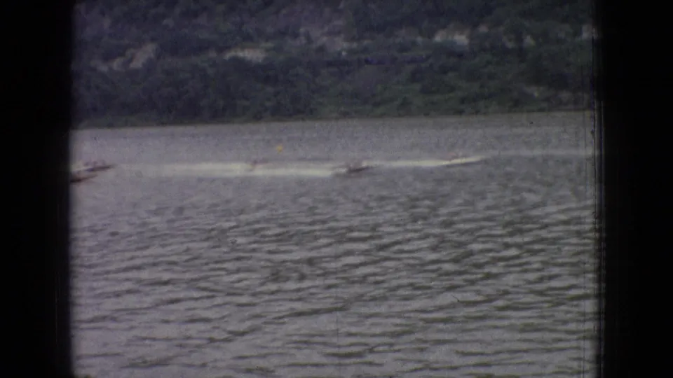 1965 New Jersey 8mm home-movie frame showing small speed boats racing across a calm lake with white wakes and a wooded far shore.