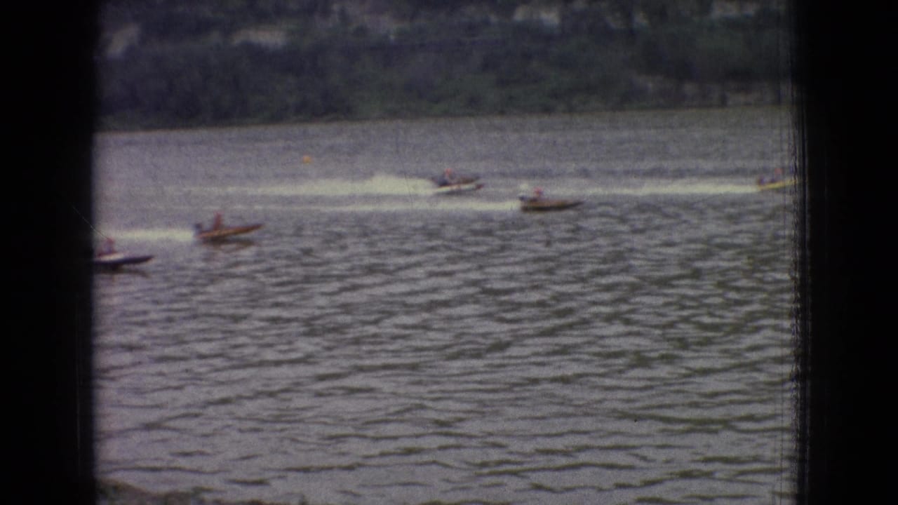 Small racing motorboats move across a New Jersey lake in 1965, seen through dark foreground bars with rippled water and distant shoreline behind them.