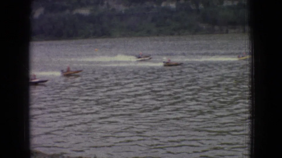 Small racing motorboats move across a New Jersey lake in 1965, seen through dark foreground bars with rippled water and distant shoreline behind them.
