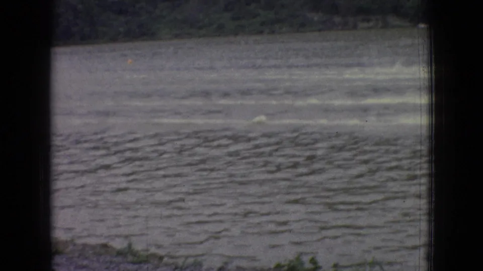A 1965 New Jersey lake view shows fading boat wakes crossing the water, with the boats small or partly out of frame and dark home-movie framing in the foreground.