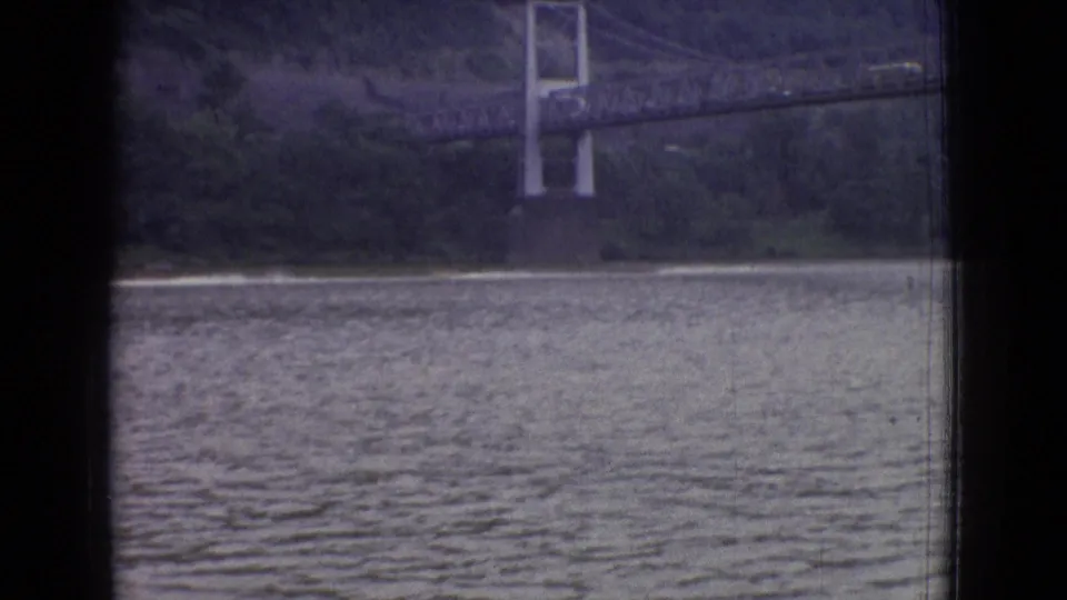 1965 New Jersey 8mm frame with a suspension bridge crossing the far shoreline above textured water, with dark film or window edges at the sides.