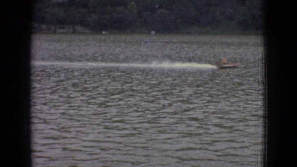 1965 New Jersey 8mm frame showing a small speedboat moving across a wide river or lake, leaving a white wake near a wooded shoreline.
